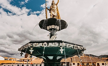 Fuente histórica en Cusco con estatua en la cima, plaza principal bajo cielo nublado